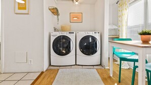 A laundry room with two front-loading washing machines, a small table with a basket, and a window with blinds. - 2 Blocks From Beach, Unit #6, Pet Friendly, Great Location, WiFi, Kitchen (Rockaway Beach)