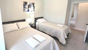Habitación de hotel con dos camas, una mesita de noche con lámpara y una puerta que da a otra habitación.