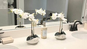 Un baño con encimeras blancas, un espejo grande, un grifo negro y tres jarrones blancos con orquídeas.
