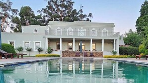 Ein Pool mit Steinbord, ein Gebäude mit überdachter Terrasse und eine Steinmauer.