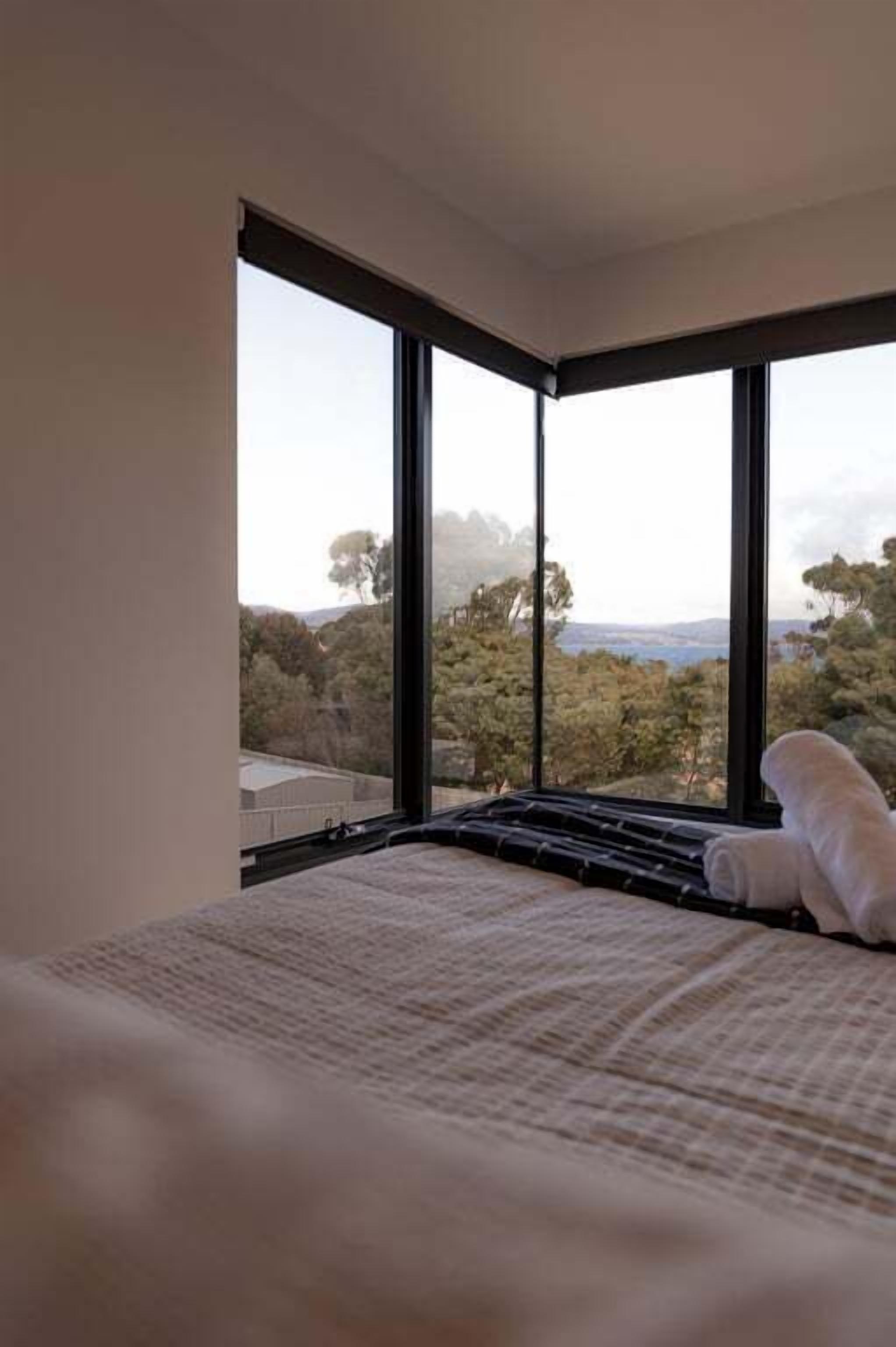 Ein Schlafzimmer mit großem Fenster, Bett und Blick auf Bäume und Hügel.