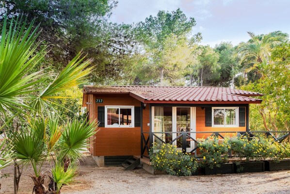 Une petite cabane en bois, surmontée d’un toit recouvert de tuiles rouges, entourée d’une végétation luxuriante et bordée d’un chemin de gravier.