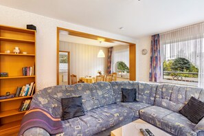 Una sala de estar con sofá, estantería, comedor y vistas a través de una ventana.