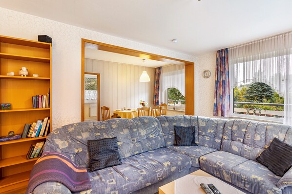 A living room with a sofa, a bookshelf, a dining area, and a view through a window. - Apartment 'Ferienwohnung Typ A' with Private Terrace and Wi-Fi (Willingen (Upland))