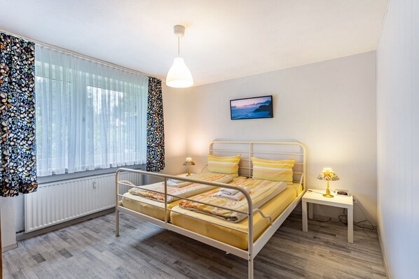 Une chambre à coucher avec un lit en métal, une table de chevet avec une lampe, une fenêtre avec des rideaux légers et un tableau accroché au mur.
