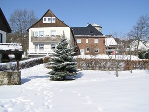Un quartier résidentiel enneigé, avec une maison à deux étages dotée d’un balcon et d’un petit sapin de Noël.