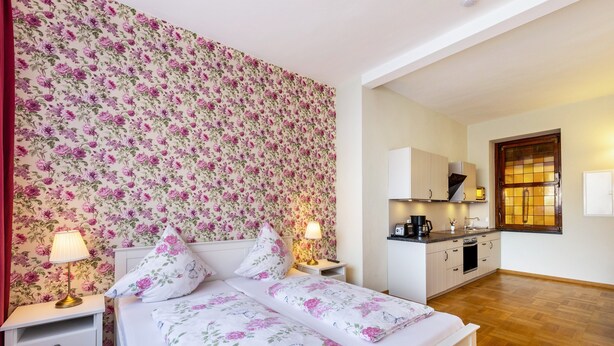 Une chambre avec un papier peint à motifs floraux, un lit double, des lampes de chevet et une vue sur la cuisine.