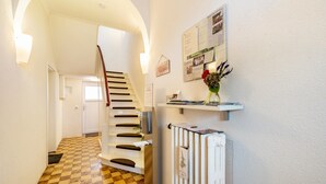 Un couloir avec un sol à carreaux, un radiateur blanc, un escalier et un tableau d’affichage recouvert de feuilles de papier.