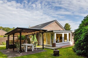 A covered patio area with a table and chairs, a brick house in the background, and a well-maintained lawn.