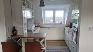 Una cocina compacta con armarios blancos, una encimera de madera y un ventanal. Hay dos taburetes de bar y una estantería para vinos.