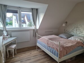 A bedroom with a sloped ceiling, a bed with a pink blanket, a bedside table with a lamp, a window with curtains, and a view of buildings outside.