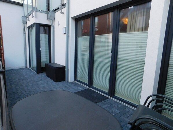Un espace patio aménagé avec une table et des chaises, un petit banc noir et une porte-fenêtre coulissante donnant sur un espace abrité.
