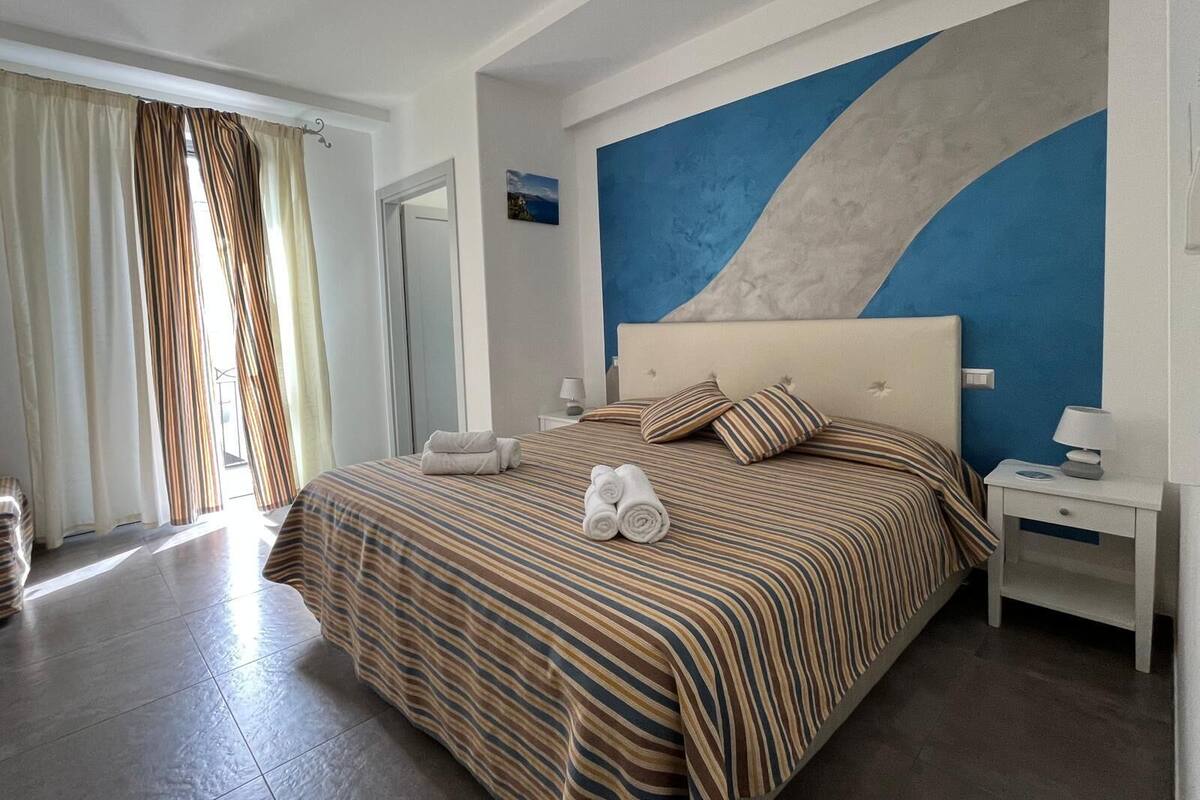 Habitación de hotel con una cama grande, una mesita de noche, una pared con diseño abstracto azul y blanco, y una ventana con cortinas.