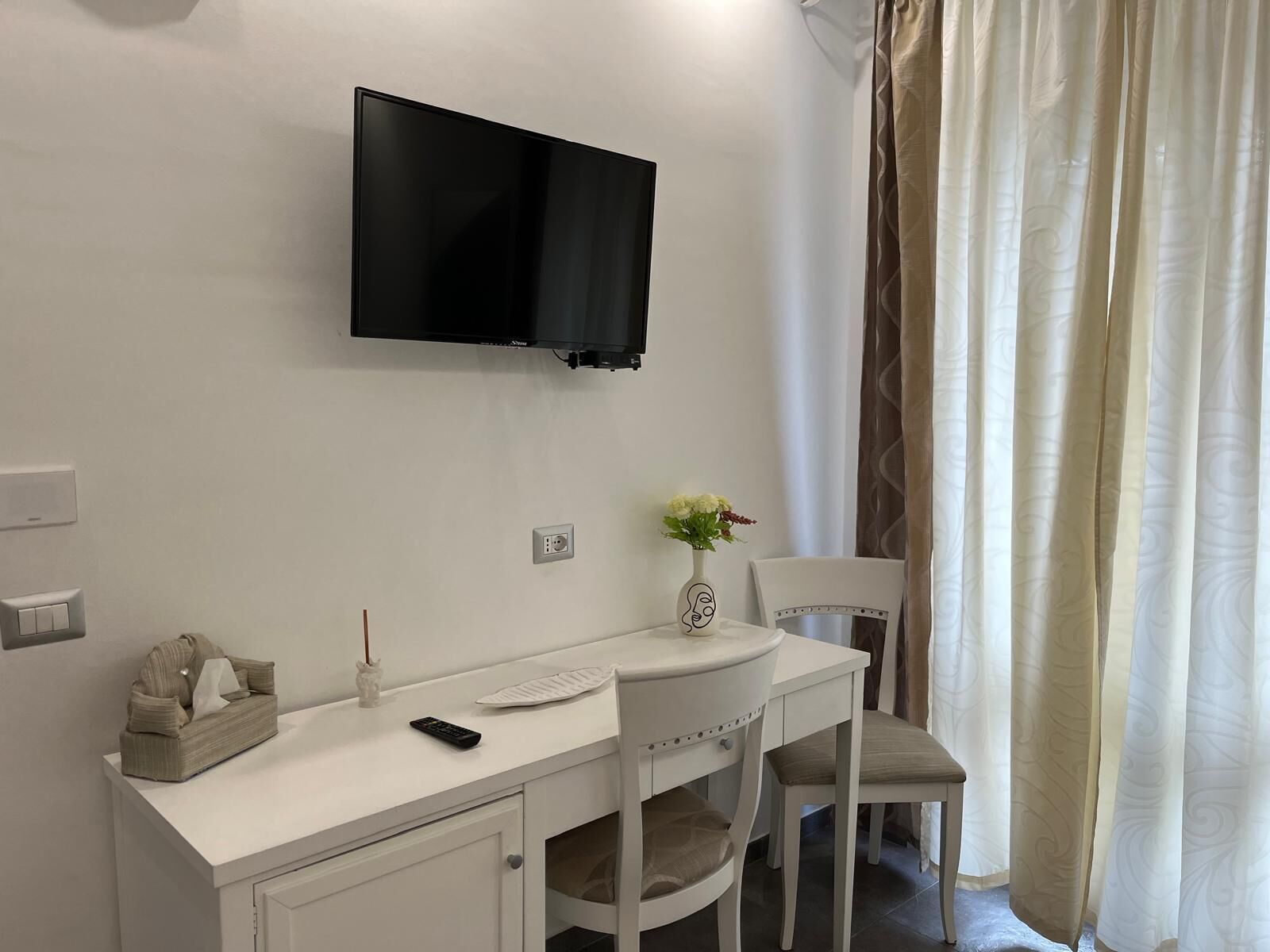Una camera d'albergo con una scrivania bianca, una sedia, una televisione a parete, un vaso con fiori e una finestra con tende.