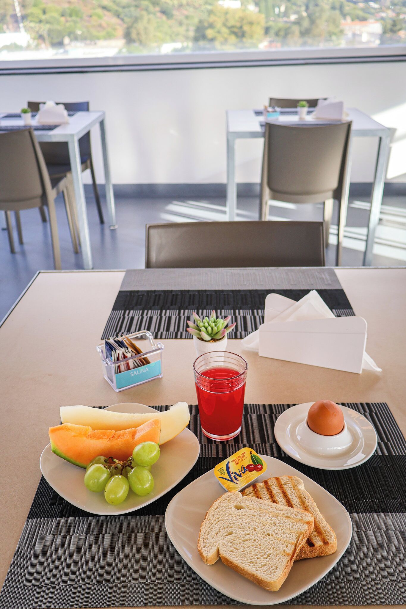 A breakfast setting with a plate of toast, a glass of red juice, a slice of melon, and a small dish of jam.