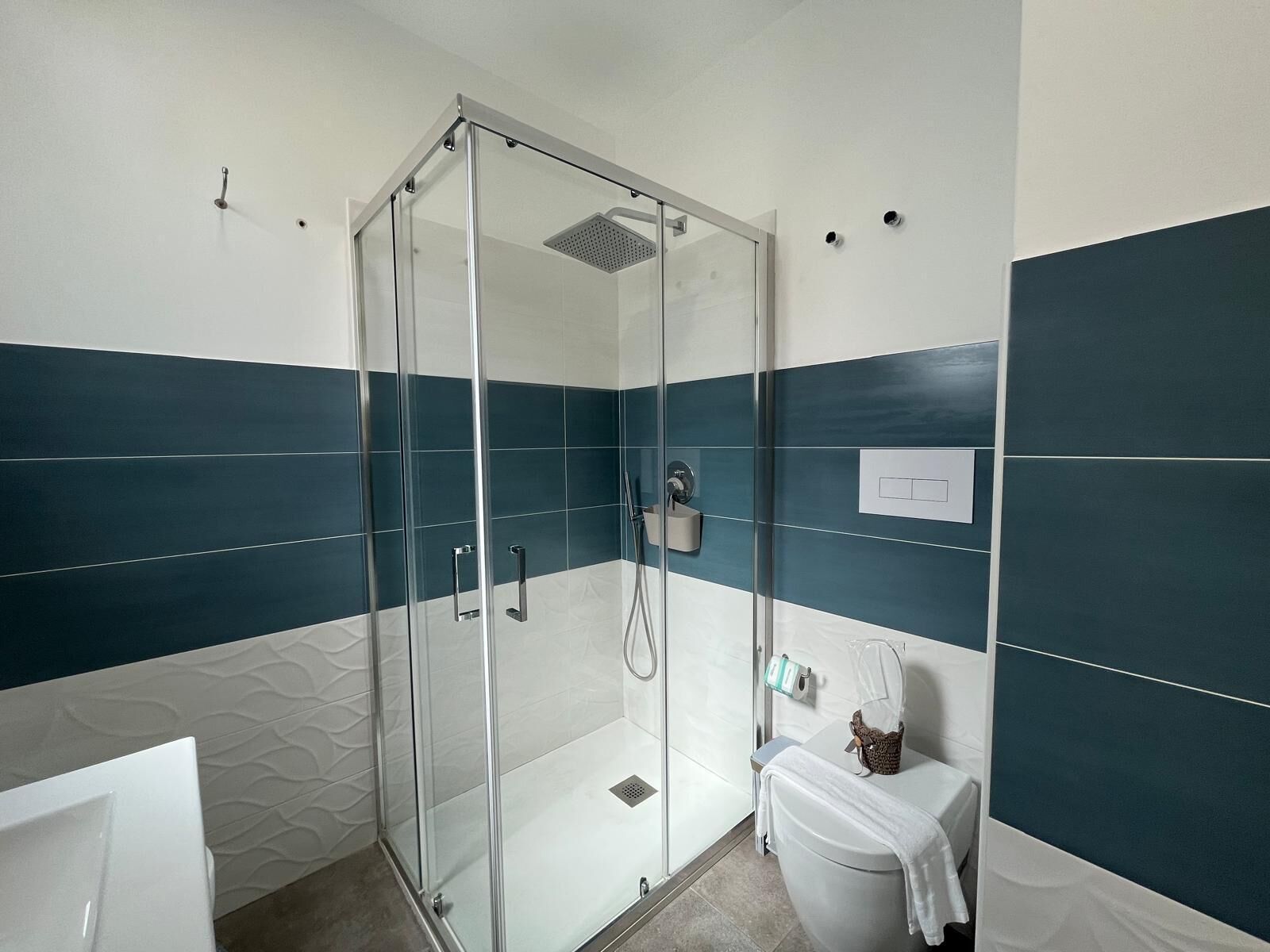 A modern bathroom with a glass-enclosed shower, white and teal tiles, a toilet, and a towel rack.