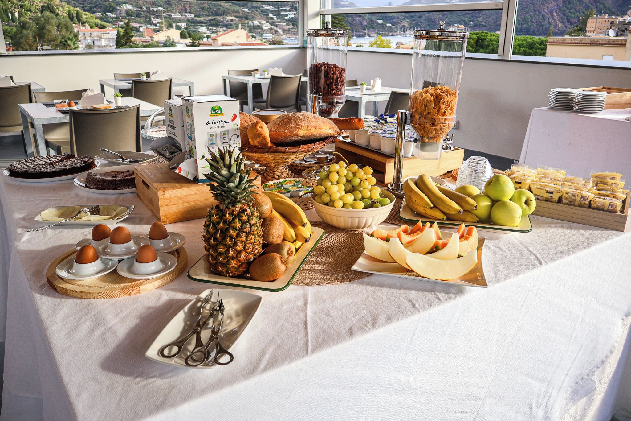 Ein Frühstücksbuffet mit verschiedenen Früchten, Brot und Gebäck auf einem Tisch, im Hintergrund ist eine Stadtansicht zu sehen.