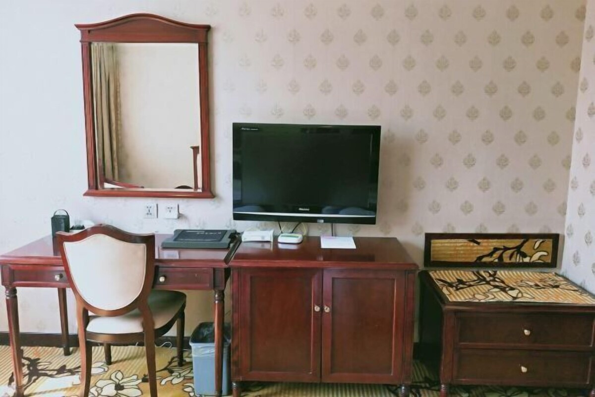 A hotel room with a wooden desk, a television, a mirror, and a bed with a patterned headboard.