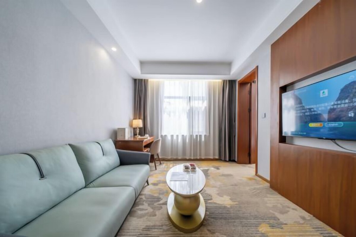 Habitación de hotel con sofá verde claro, una mesita de centro redonda, televisor montado en la pared, escritorio con lámpara y ventana con cortinas.