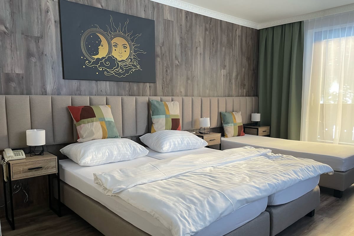 A hotel room with a large bed, two bedside tables, a nightstand, a wall-mounted artwork, and a window with curtains.