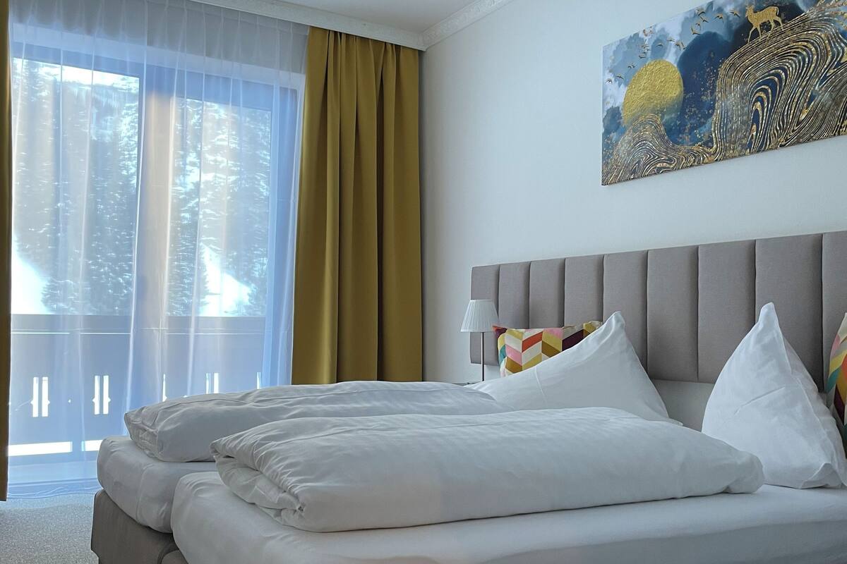 A hotel room with a large bed, a view of trees through sheer curtains, and a painting on the wall.
