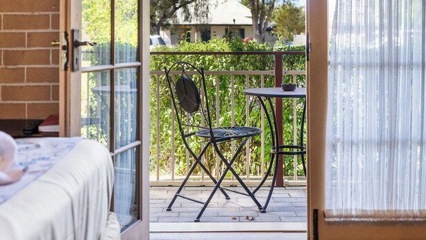 Ein Zimmer mit einem Bett, einer Glastür, die auf einen Balkon mit Tisch und Stühlen führt, und Blick auf Bäume und Gebäude.