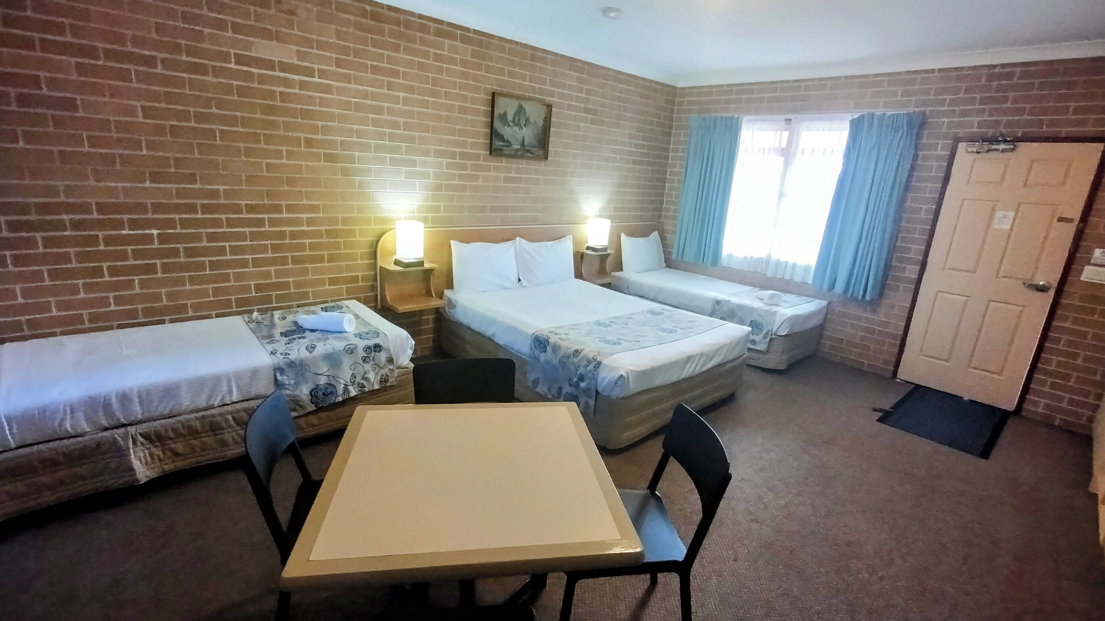 A hotel room with two single beds, a small table, and a chair. There is a brick wall, a painting, and a door.