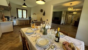 A dining area with a table set for four, including plates, cutlery, and a bottle of wine. There are decorative items like flowers and a vase on the table. The room has a modern kitchen in the background with a dining area visible through an open door. - House in Tuscany near Pisa & Lucca (Lucca)