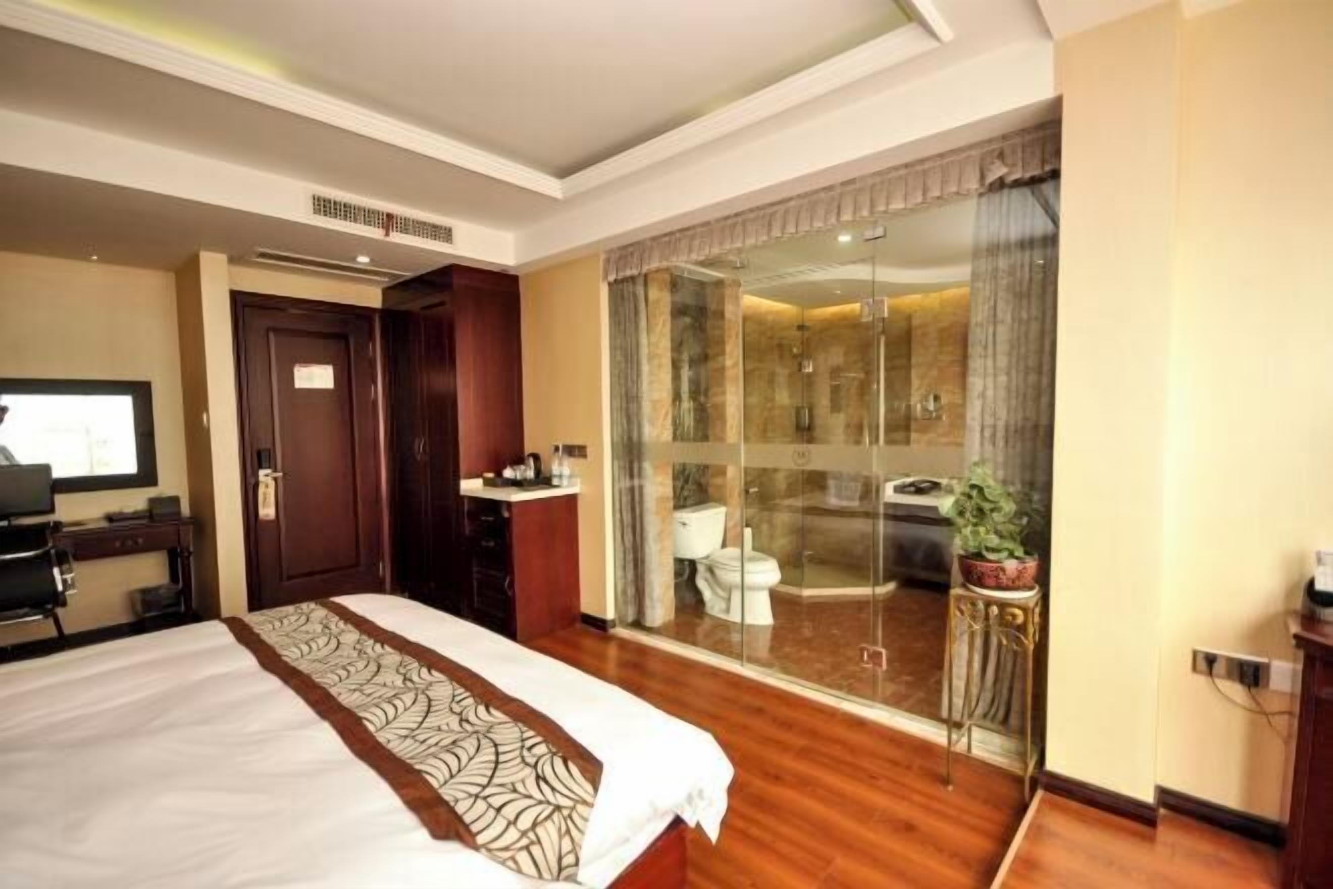 A hotel room with a bed, a desk, a bathroom with a shower, and a glass door leading to a balcony.