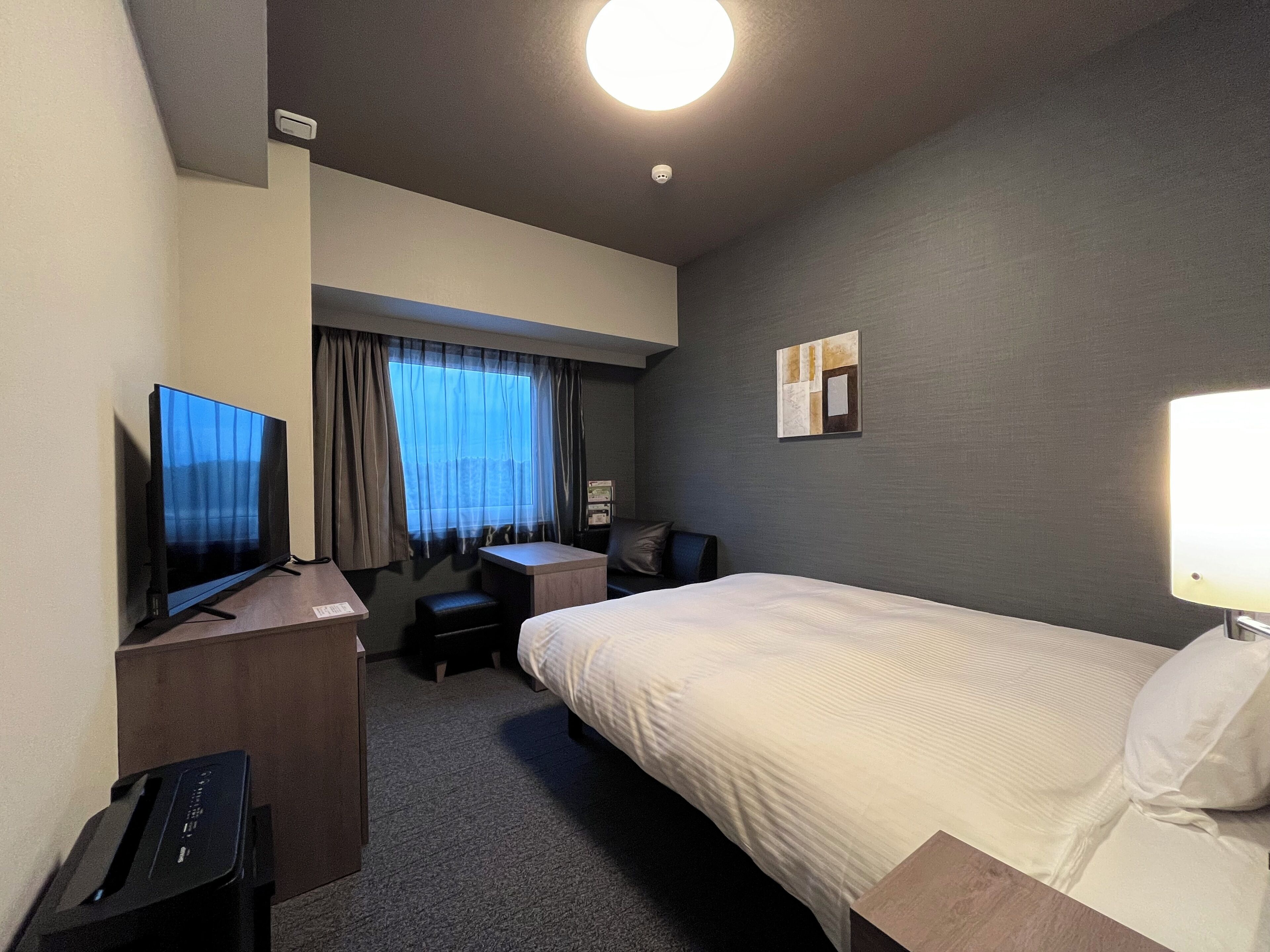A hotel room with a bed, a desk with a computer, a TV, a chair, a small table, and a window with curtains.