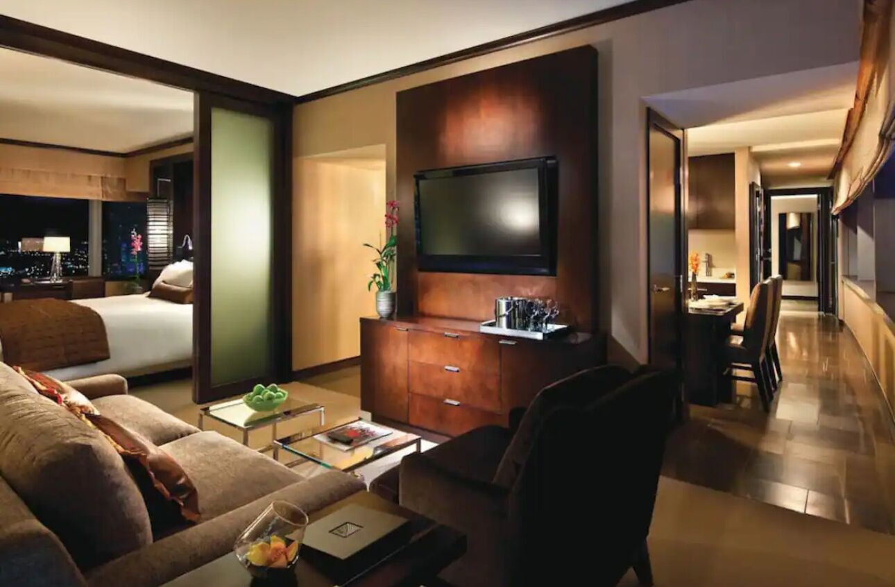 A hotel room with a large bed, a TV mounted on a wooden cabinet, a seating area with a coffee table, and a view of the city at night.