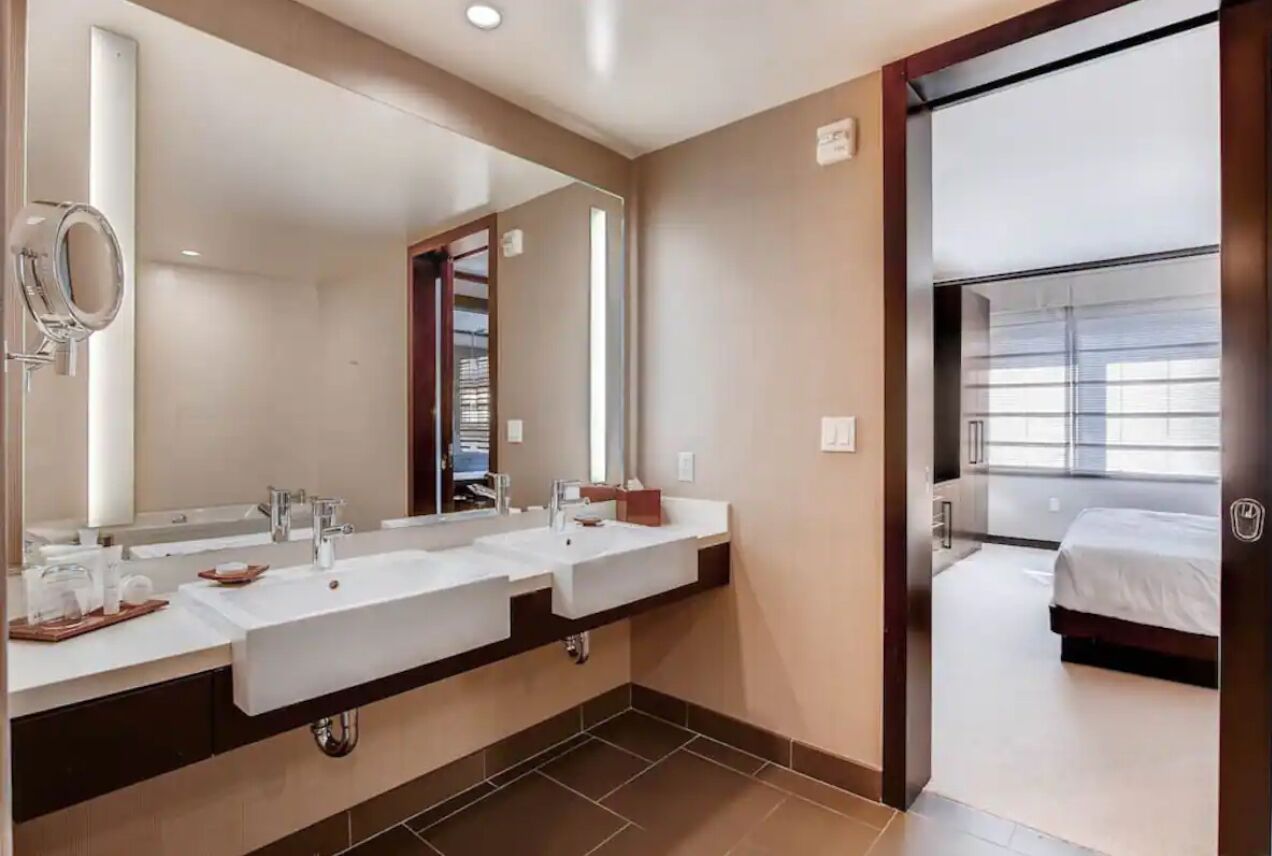 A modern bathroom with two sinks, a large mirror, and a view of a bedroom through an open door.