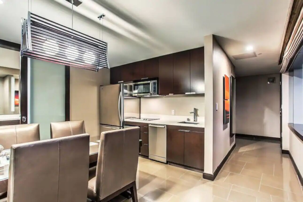 Una cocina moderna con armarios oscuros, un refrigerador de acero inoxidable y un microondas. Cuenta con una zona de comedor con sillas de cuero y una mesa.