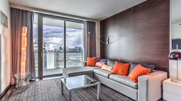 A modern hotel room with a large sliding glass door leading to a balcony, a grey sofa with orange cushions, a glass coffee table, and a dark wood accent wall. - Studios With Gorgeous Views Of The Strip! (Las Vegas)