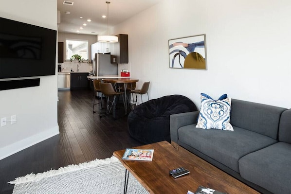 Un salon moderne avec un canapé gris, un fauteuil en forme de poire noir et une table basse en bois. On aperçoit à l’arrière-plan un coin repas avec une table et des chaises.