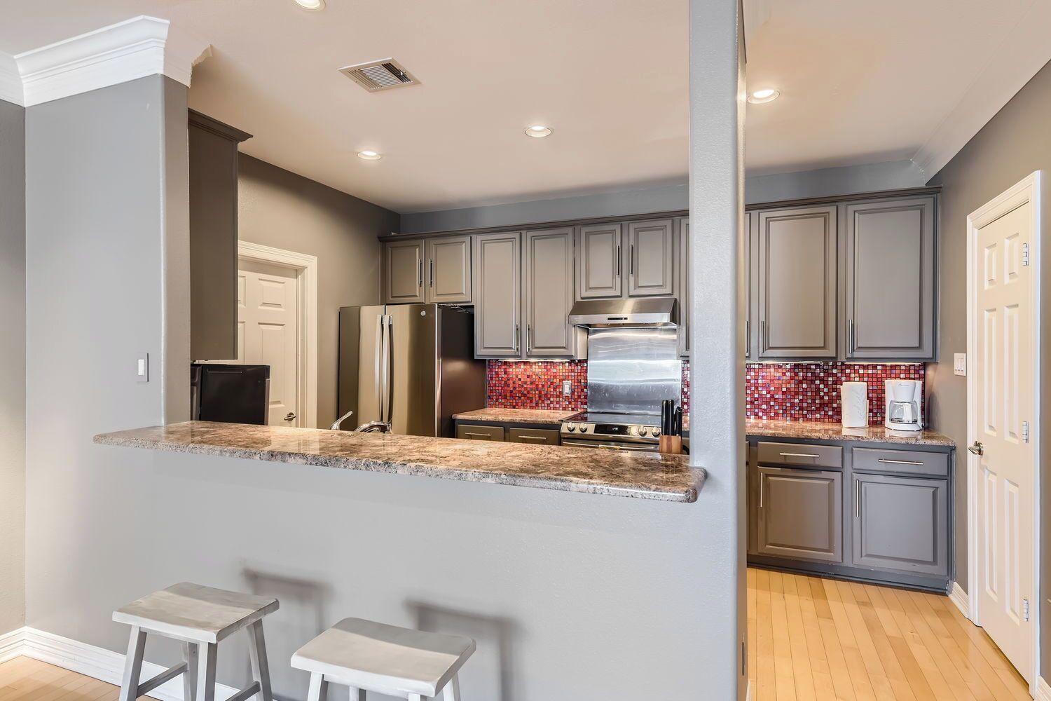 A modern kitchen with grey cabinets, a stainless steel refrigerator, a stove, and a coffee maker. There are two bar stools and a red mosaic backsplash.