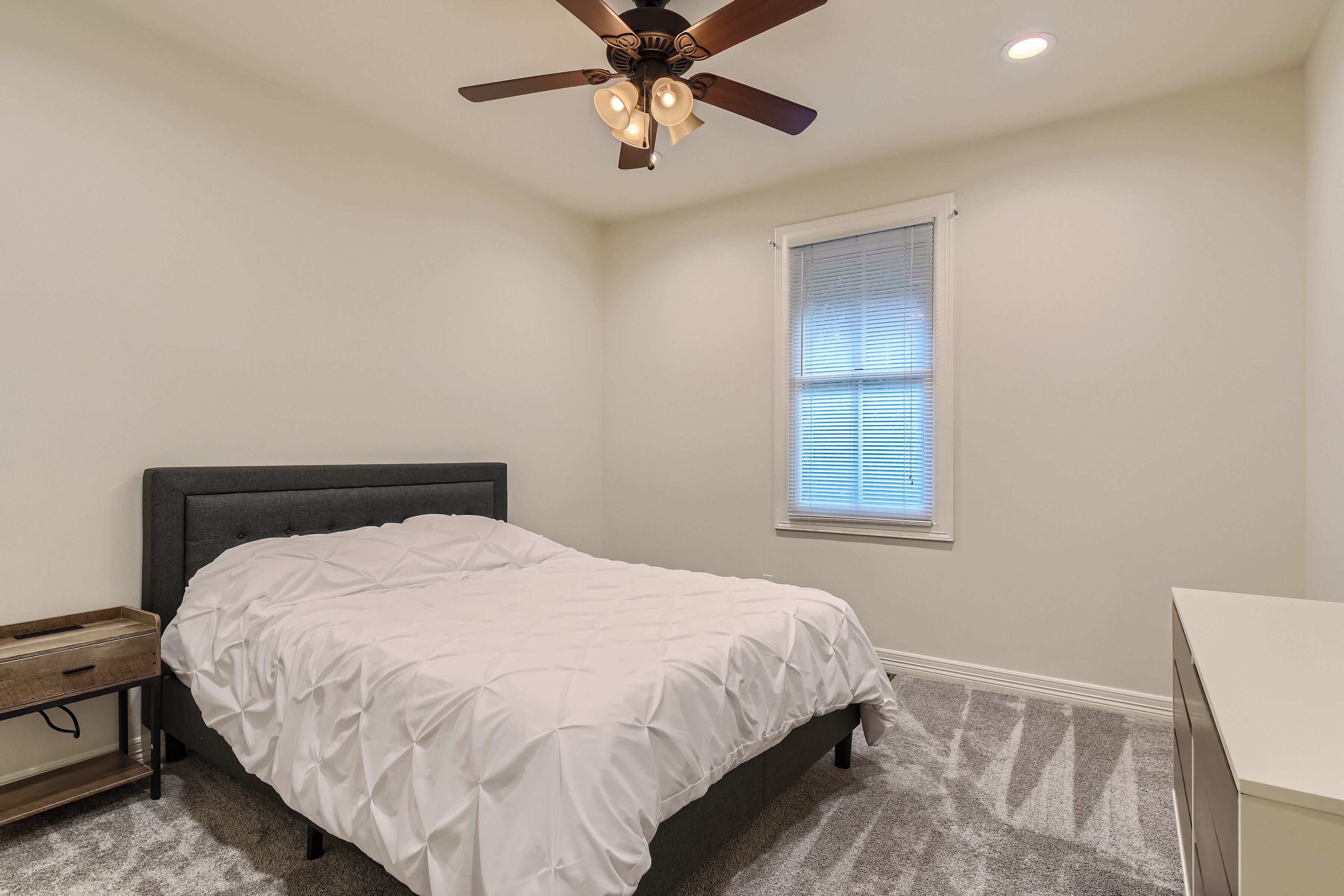 A bedroom with a bed, a nightstand, a desk, a ceiling fan, and a window with blinds.