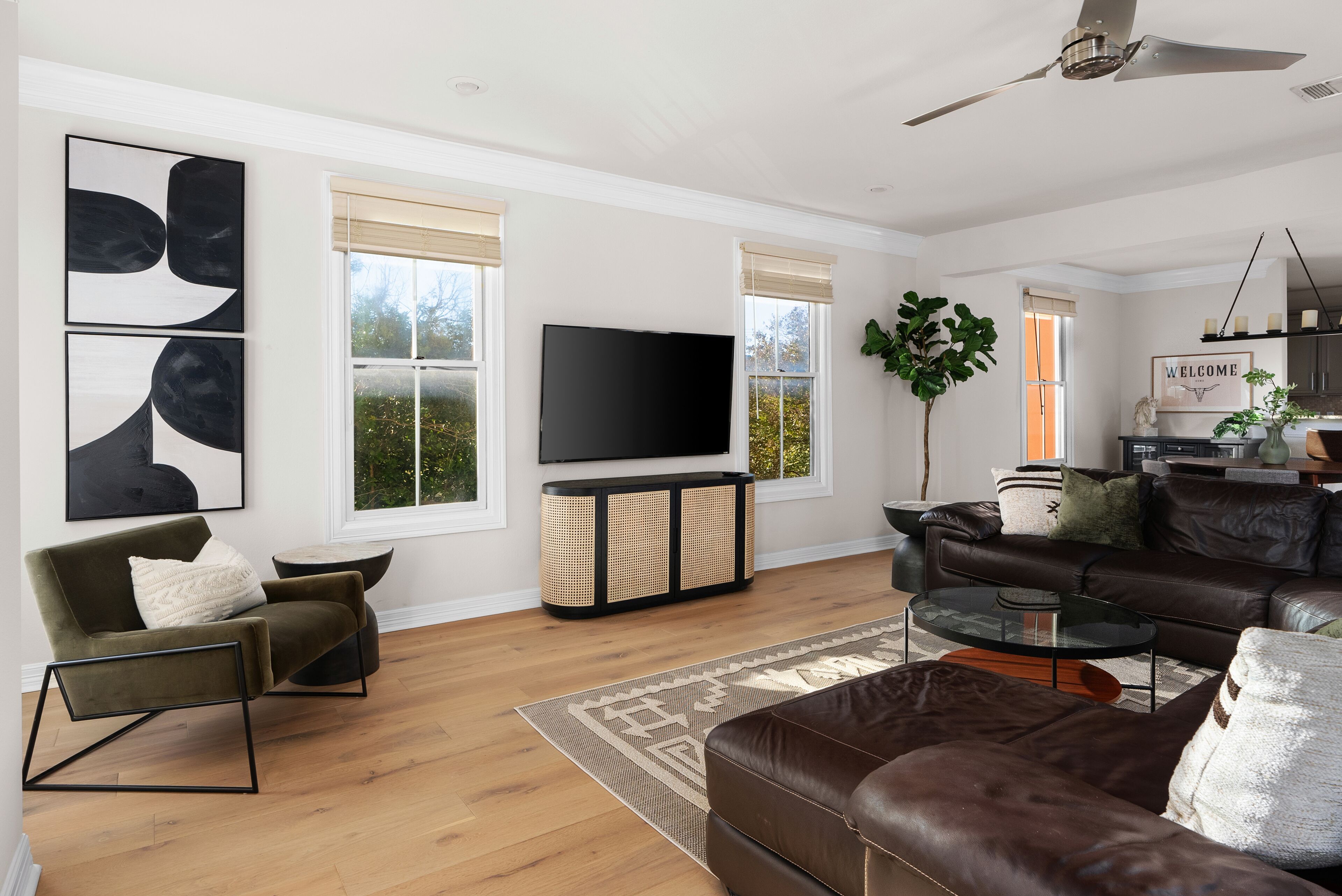A modern living room with a sofa, armchair, and coffee table. There is a flat-screen TV on a stand, a plant, and a window with blinds.