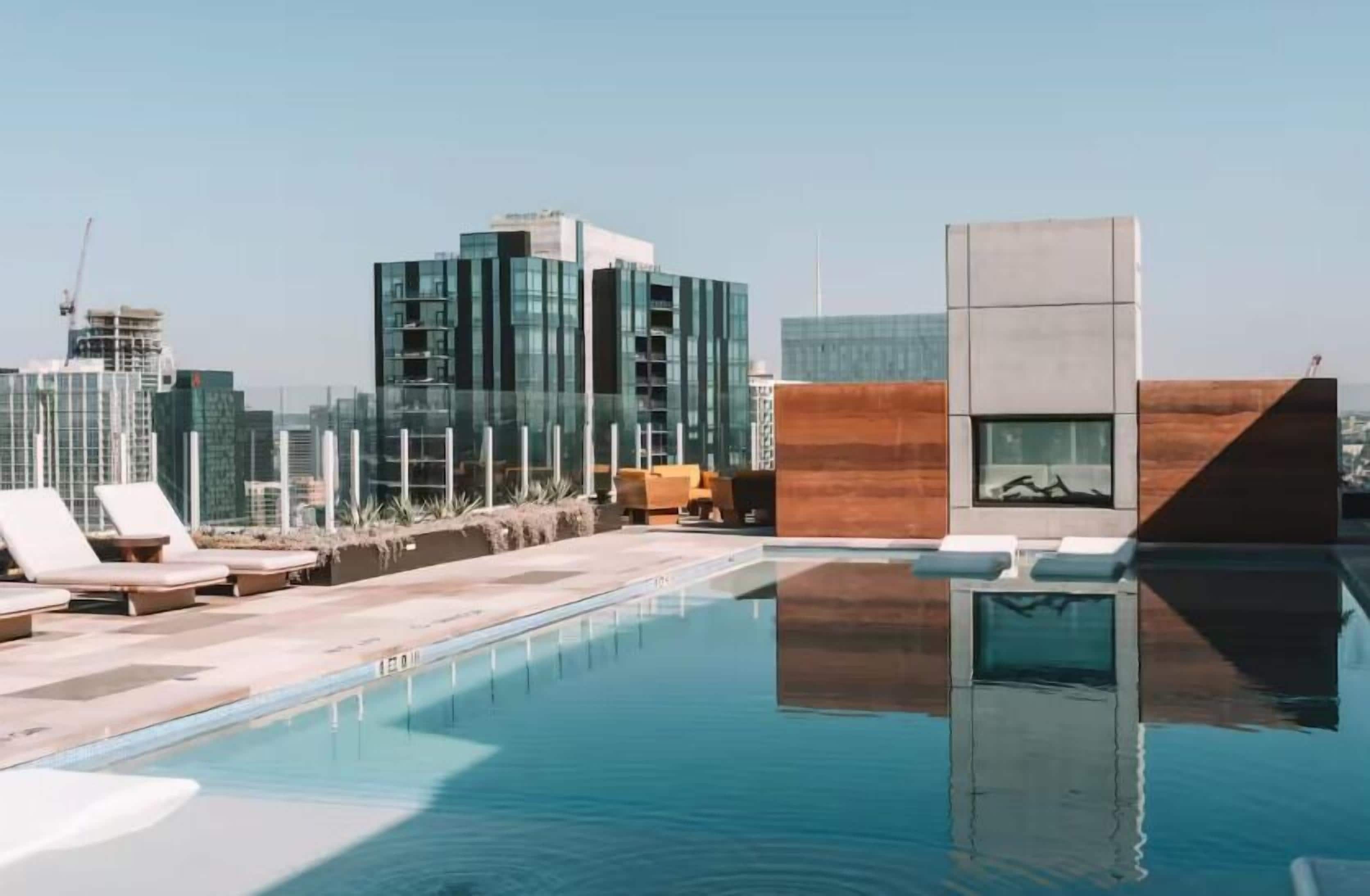 A rooftop pool with lounge chairs, a wooden structure, and a city skyline in the background.