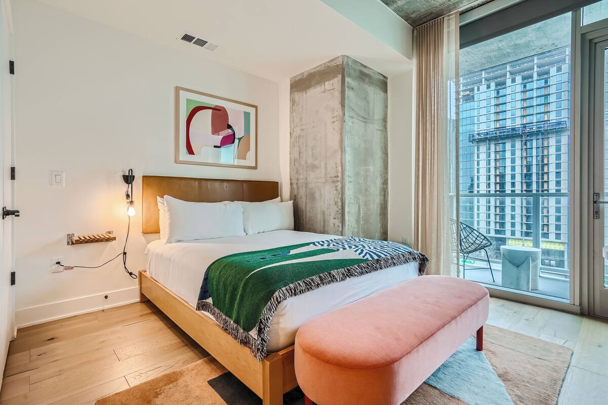 Un dormitorio moderno con una cama de madera, una manta estampada en verde y blanco, un banco rosa y un ventanal con vistas a la ciudad.