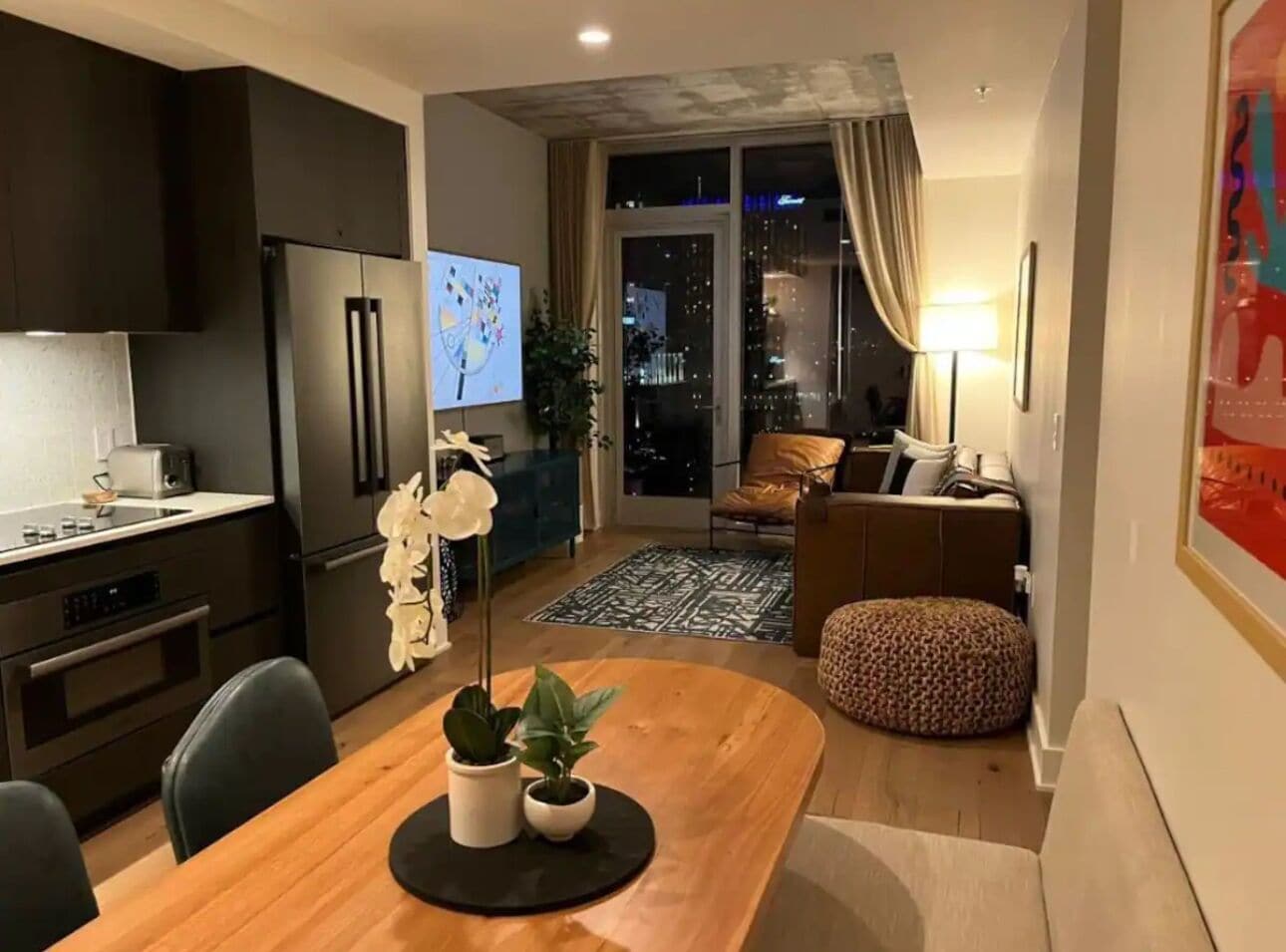 Une cuisine moderne aux meubles sombres, un coin repas avec une table et des chaises en bois, et un salon avec un canapé et un pouf.