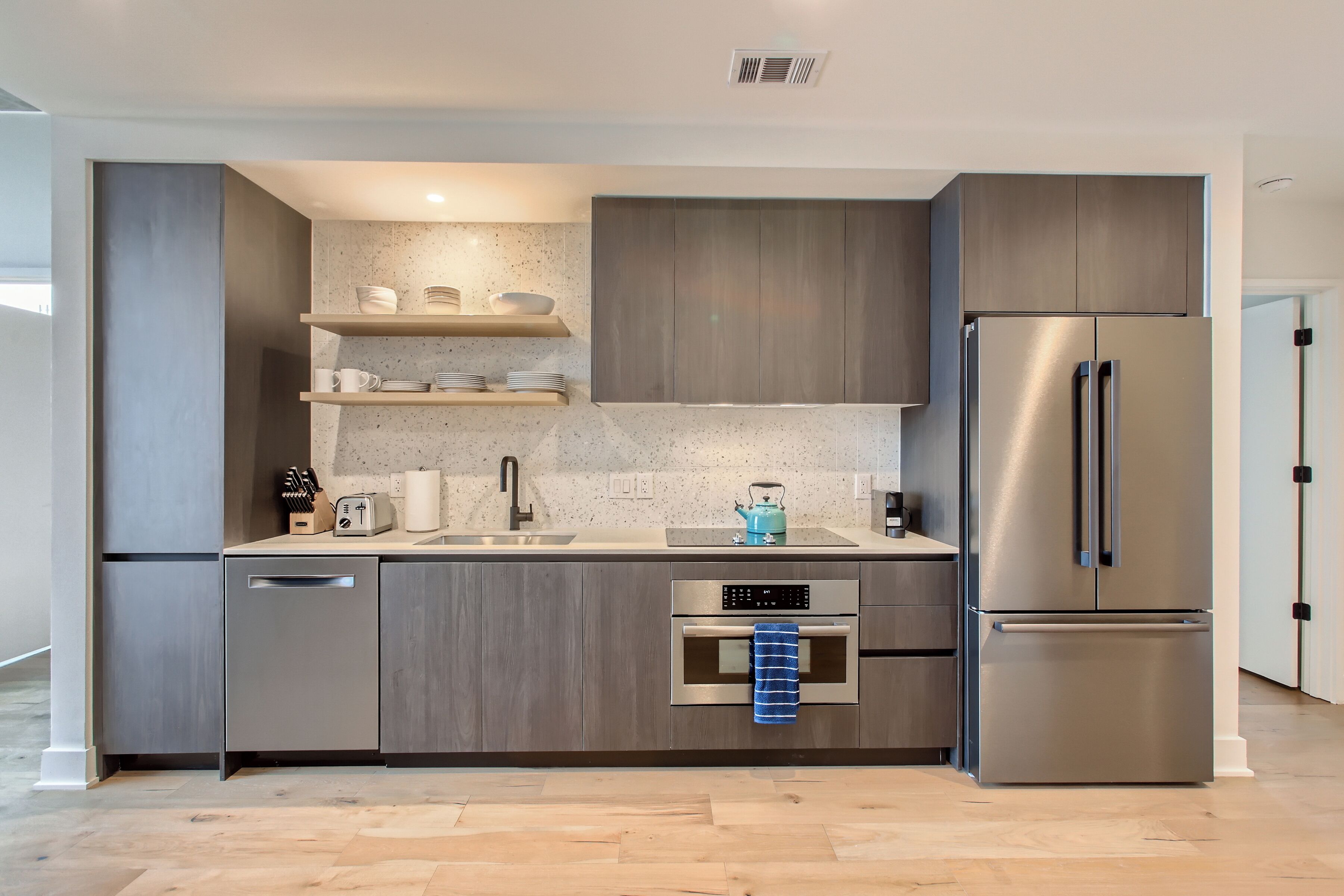 A modern kitchen with grey cabinetry, a stainless steel refrigerator, a built-in oven, and a countertop with a kettle and utensils.