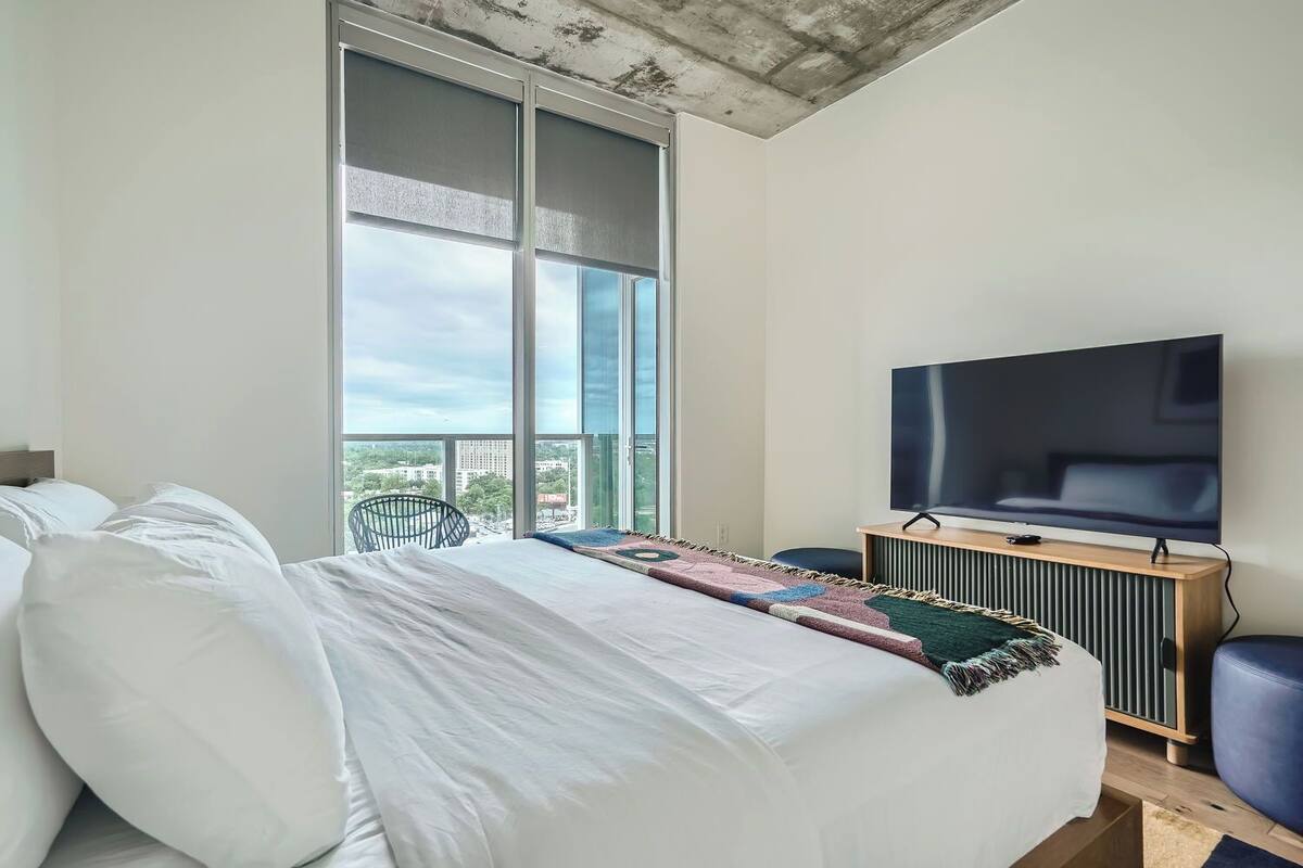 Un dormitorio moderno con una cama grande, un televisor y vistas a la ciudad a través de amplios ventanales.