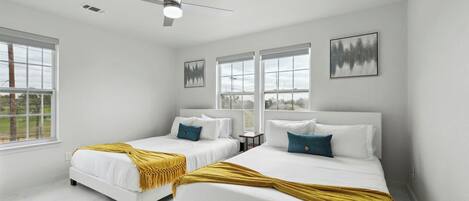 A hotel room with two beds, white bedding, a ceiling fan, large windows, and artwork on the walls.