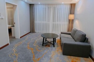 Una habitación de hotel moderna con un sofá gris, una mesa redonda y un ventanal con cortinas translúcidas.