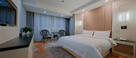 A hotel room with a bed, a desk, a chair, a small table, and a potted plant.
