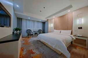 Quarto de hotel com cama, escrivaninha, cadeira, mesa pequena e planta em vaso.