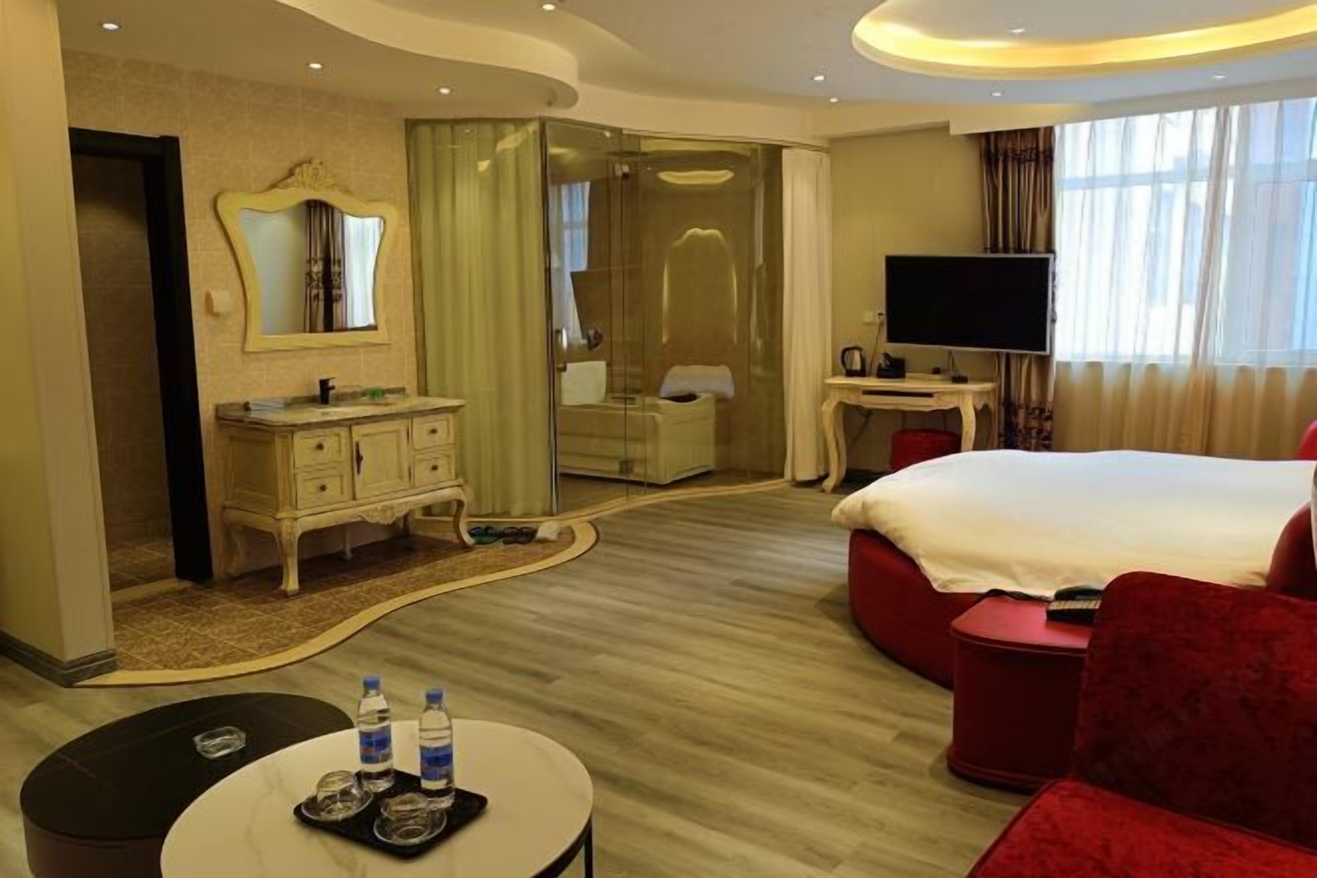 A hotel room with a bed, a TV, a desk, a mirror, a vanity, and a shower.