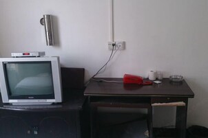 A room with a television on a stand, a desk with a roll of toilet paper, a glass ashtray, and a wall-mounted heater.