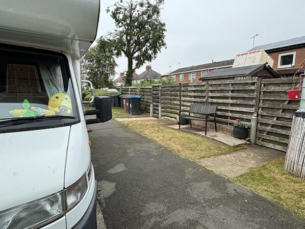 A residential driveway with a white van parked on the left. There is a wooden fence on the right, and a bench is visible in the background. - Motorhome in Hinckley- Sleeps 3- Pet Friendly (Hinckley)